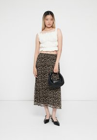 Frau in einem weißen, gerafften Crop-Top, einem schwarz-beige gemusterten Midi-Rock, schwarzen High Heels, die eine schwarze Handtasche vor einem weißen Hintergrund hält.