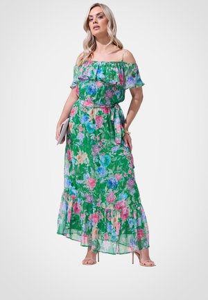 Kobieta w zielonej, kwiecistej sukience maxi z odkrytymi ramionami, trzymająca kopertówkę, stojąca na białym tle w cielistych sandałach na wysokim obcasie.