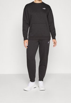 Persona con sudadera negra de Puma y pantalones deportivos, con zapatillas blancas, de pie sobre fondo claro y liso.