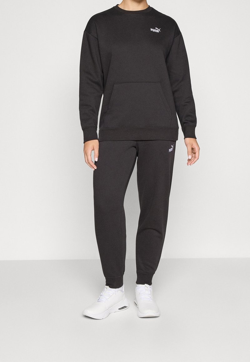 Persoon met een zwarte Puma-trui en joggers, witte sneakers, staand tegen een egale lichte achtergrond.