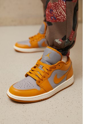 Par orange og grå Nike Air Jordan sneakers brugt med brune bukser med røde og sorte grafiske tryk, stående på et lyst plettet gulv.