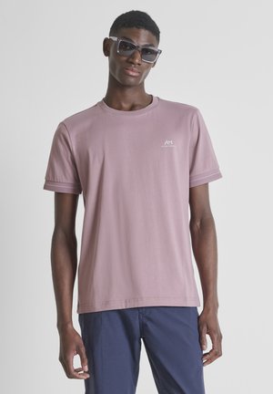 Giovane uomo con grandi occhiali da sole scuri, maglietta a maniche corte color malva chiaro con piccolo logo e pantaloni blu navy, in piedi davanti a uno sfondo semplice.