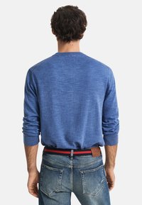 Man med lockigt mörkt hår iklädd en blå långärmad skjorta instoppad i urtvättade jeans med ett marinblått och rött bälte, vänd bort.