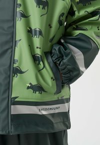Enfant portant une veste de pluie verte à motifs de dinosaures avec des bandes réfléchissantes et la marque "Swedemount", main dans la poche.