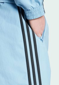 Light blue athletic jacket featuring black stripes along the sides and a zippered pocket. Material appears smooth with elastic cuffs.