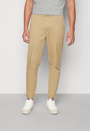 Tan chinos dengan desain ramping, pinggang elastis, saku samping, dan manset, dipadukan dengan sepatu sneakers putih yang memiliki sol bertekstur.