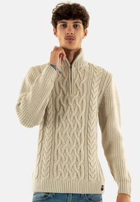 Crème kabelgebreide trui met een hoge kraag en een halve rits, met textuurpatronen en een geribbelde zoom en manchetten.