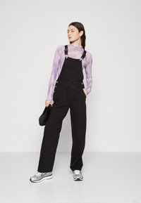 Black denim overalls with adjustable straps, side buttons, and a loose fit, worn over a sheer purple long-sleeve top; styled with silver sneakers.