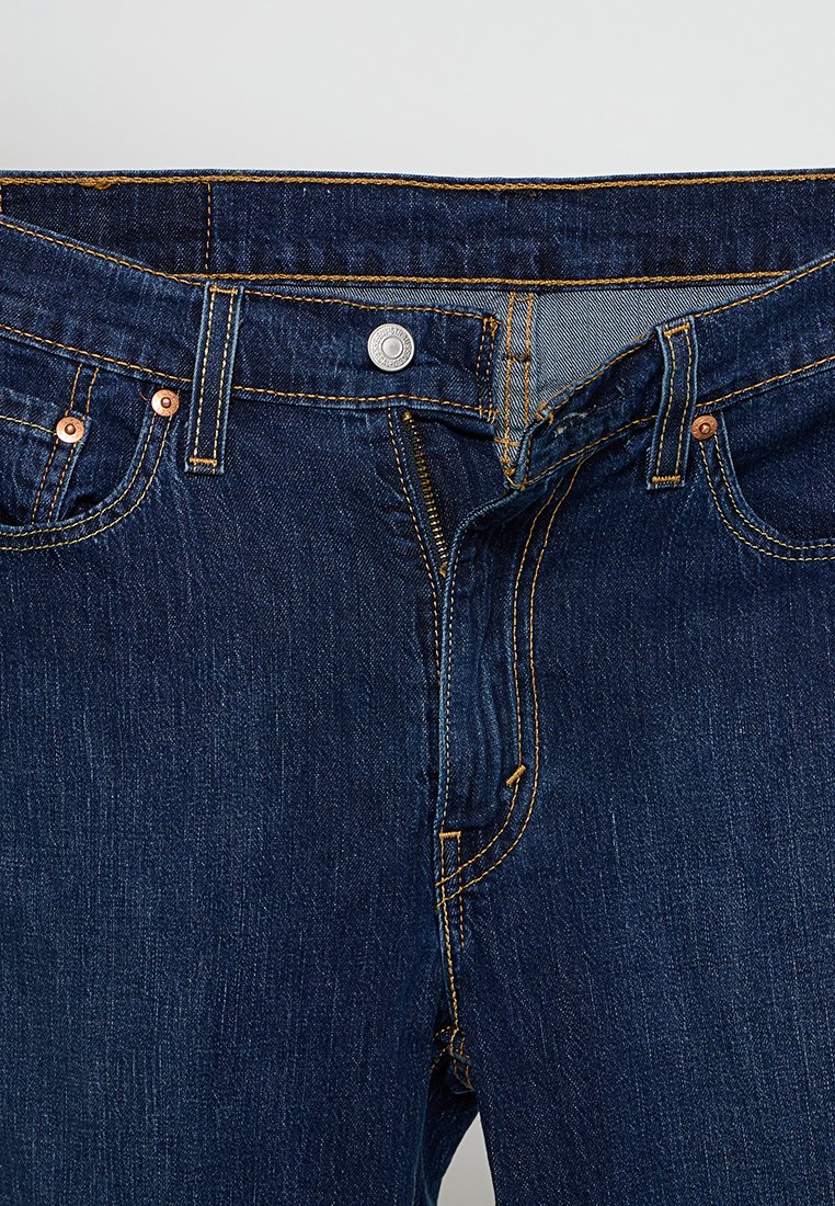 Dunkelblaue Denim-Jeans mit Kupferbeschlägen, gelber Naht, Reißverschluss und fünf Taschen; mittlere Taille und gerade Beine.
