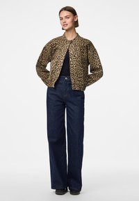 Leopardmönstrad bomberjacka med elastiska ärmar, kombinerad med högmidjade mörkblå jeans och svarta skor på en modell mot en ljus bakgrund.