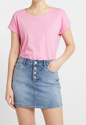 Kobieta ubrana w różowy krótki t-shirt włożony do niebieskiej dżinsowej spódnicy z wysokim stanem i guzikami z przodu, z rękami częściowo w kieszeniach.