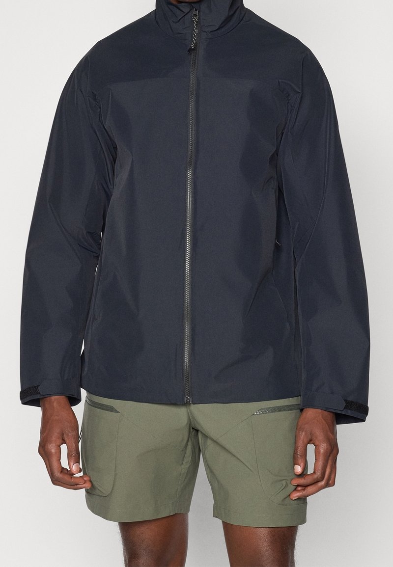Veste imperméable navy avec col haut et fermeture éclair sur toute la longueur. Présente des poignets élastiques et deux poches latérales zippées. Assorti avec un short olive.