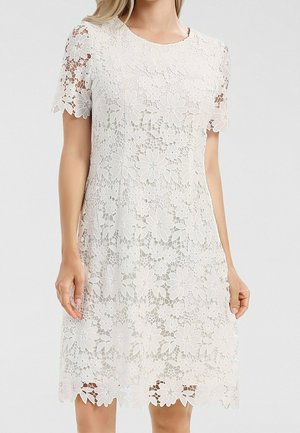 Femme portant une robe blanche en dentelle mi-longue avec des manches courtes et des motifs floraux sur une doublure unie.