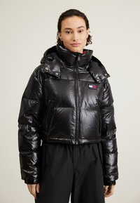 Chaqueta pimienta negra con acabado brillante, diseño corto, cuello alto, dos bolsillos frontales y un parche de logo en el pecho. Con capucha.