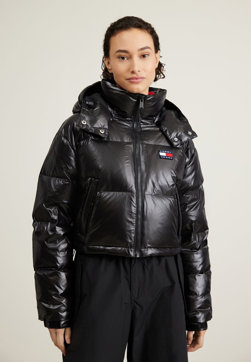 Chaqueta pimienta negra con acabado brillante, diseño corto, cuello alto, dos bolsillos frontales y un parche de logo en el pecho. Con capucha.