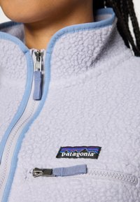 Tuvas tuvuma gaisā purpursarkana flīsa jaka ar zilu apdari, aizdara, ar Patagonia logo uzlīmi un rāvējslēdzēja krūšu kabatu.