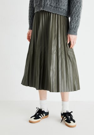 Gonna plissettata lunga fino alla caviglia di colore verde oliva, realizzata in ecopelle. Abbbinata a sneaker nere, calzini bianchi a girocollo e un maglione grigio a trama.