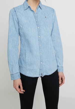 Lyseblå denimskjorte med knap-ned design, lange ærmer og et lille logo på brystet. Har stitched detaljer og en buet kant.