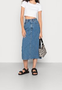 Jeanskjol, med längd till mitten av vaden, blå tvätt, framknäppning, tillsammans med en vit croppad t-shirt och svarta sandaler, där en väska med zebramönster hålls.