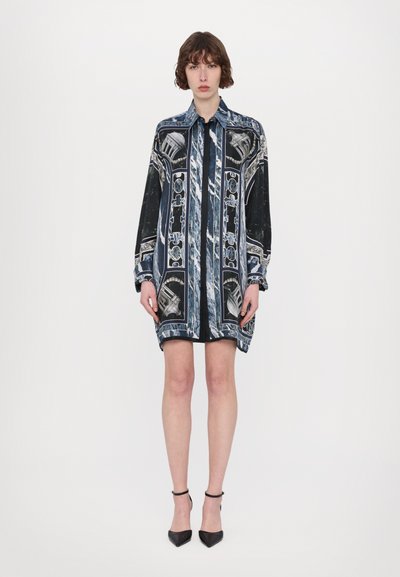 Hemdblusenkleid aus blauem und schwarzem Stoff mit filigranen Mustern, langen Ärmeln, Knopfverschluss und geradem Schnitt. Abgerundet mit spitzen schwarzen Schuhen.