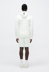 Bela športna jopa z navpičnimi črnimi črtami na hrbtu, kombinirana z usklajenimi kratkimi hlačami, obuta z belimi nogavicami in svetlimi supergami.