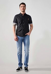 Hombre que lleva puesta una camisa negra de manga corta con botones, jeans de color azul claro y zapatillas negras, de pie con una mano en el bolsillo frente a un fondo plano.