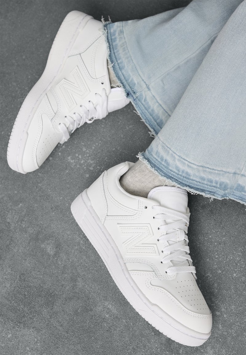 Baskets blanches avec logo "N", portées avec des chaussettes gris clair et un jean bleu clair à ourlet effiloché sur un sol gris texturé.