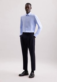 Camicia azzurro chiaro con bottoni e colletto, abbinata a pantaloni scuri su misura e scarpe di pelle nere. Tessuto liscio e design aderente.