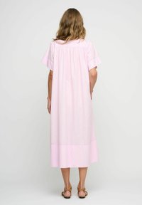 Abito di cotone rosa con maniche corte, lungo fino al ginocchio, presenta un design con schiena arricciata e orlo inferiore semplice. Indossato con sandali marroni con cinturini.