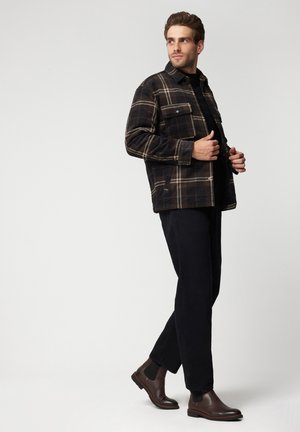 Dark plaid shirt jacket with cream accents, paired with black trousers and brown chelsea boots. The jacket features two chest pockets.
