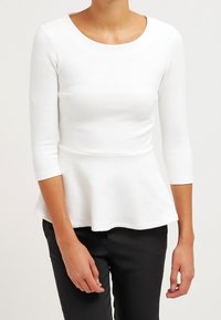 Weißes Peplum-Top mit Dreiviertelärmeln, strukturiertem Stoff, Rundhals und taillierter Passform, kombiniert mit schwarzen Hosen.