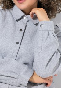 Grijze gevoerde jacket met een kraag, grote knopen als sluiting en geplooide mouwen. Zachte textuur en een relaxed fit benadrukt.