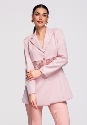 Žena v světle růžovém saku s krajkovým středem a odpovídajícími kalhotami, stojící s překříženýma rukama před světlým pozadím.