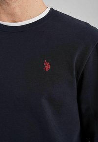 Marinblå långärmad skjorta med rund halsringning och vit innerkrage. Har en liten röd broderad logotyp på bröstet.