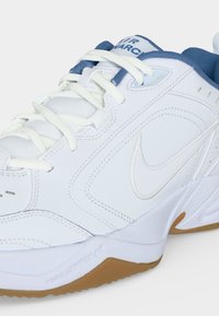Λευκό αθλητικό παπούτσι Nike Air Monarch με μπλε εσωτερική επένδυση, λευκά κορδόνια και καφέ λαστιχένια σόλα, που δείχνει λεπτομέρειες από το πλάι και το εμπρός μέρος.