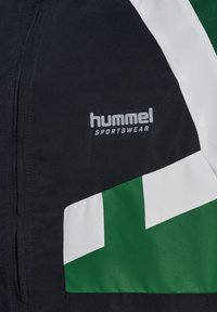 Črn športni jopič z zelenimi in belimi geometrijskimi vzorci, s sivo logo "hummel SPORTSWEAR" v ospredju. Gladka tekstura.