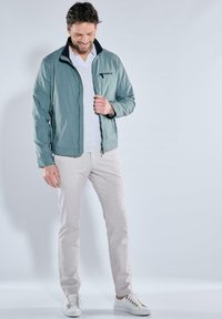 Hellgrüne Reißverschlussjacke mit schwarzem Kragen, grauem T-Shirt, hellgrauer Hose und weißen Turnschuhen; glatte Textur, lässiges Design, keine Muster sichtbar.