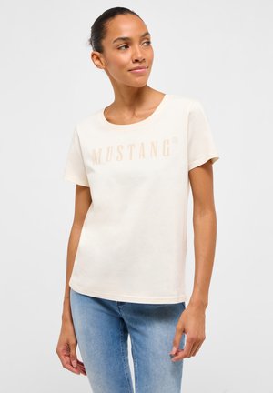 Femme portant un t-shirt Mustang crème et un jean bleu, regardant sur le côté avec les mains détendues le long du corps, sur un fond uni.