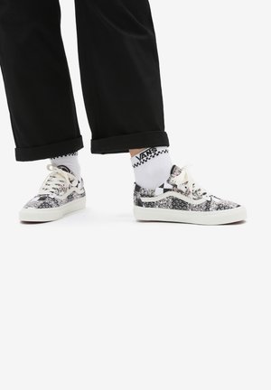 Person iført sorte bukser, hvide Vans sokker og sort-hvide Vans sneakers med blomsterprint og snørebånd, stående på hvid baggrund.