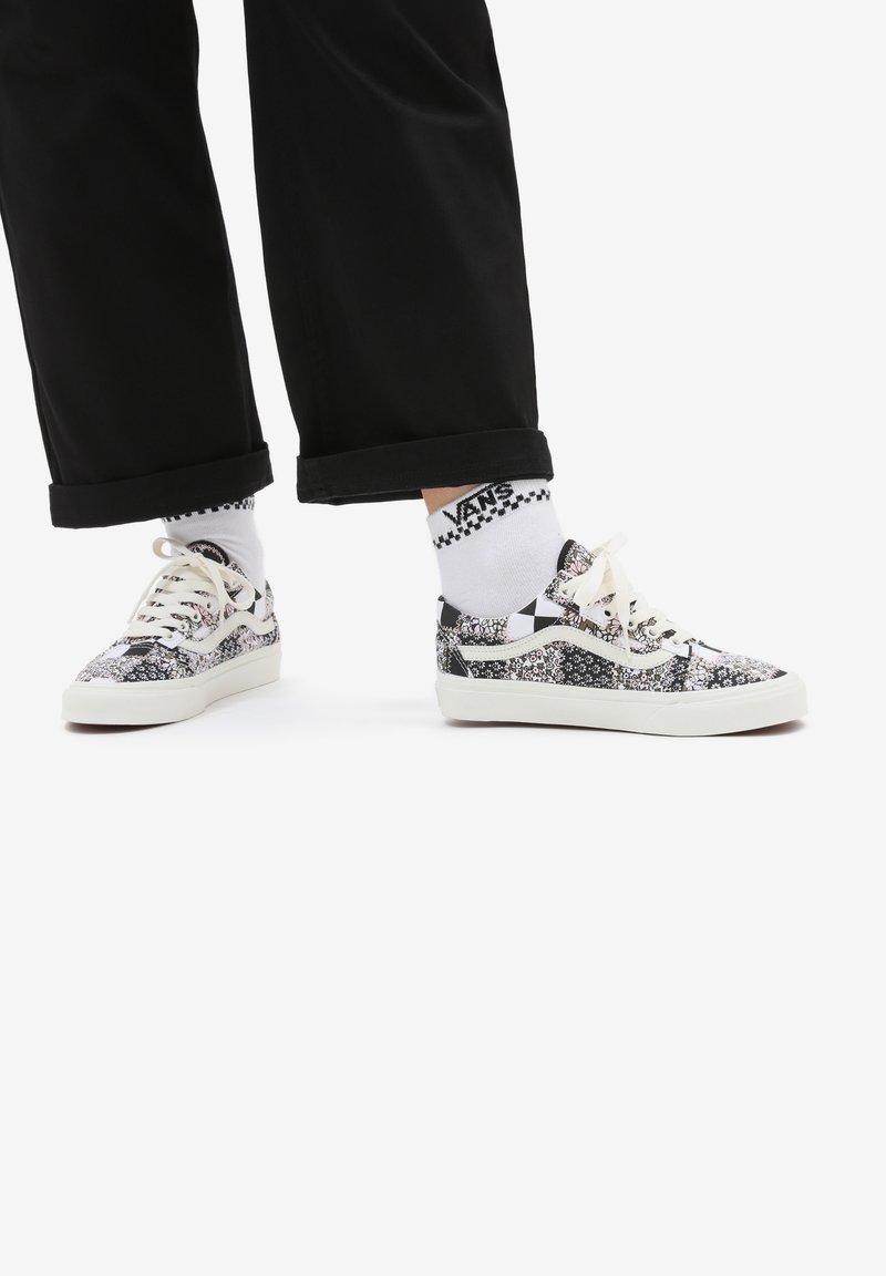 Personne portant un pantalon noir, des chaussettes blanches Vans, et des baskets Vans noire et blanche à motif floral avec des lacets, debout sur fond blanc.