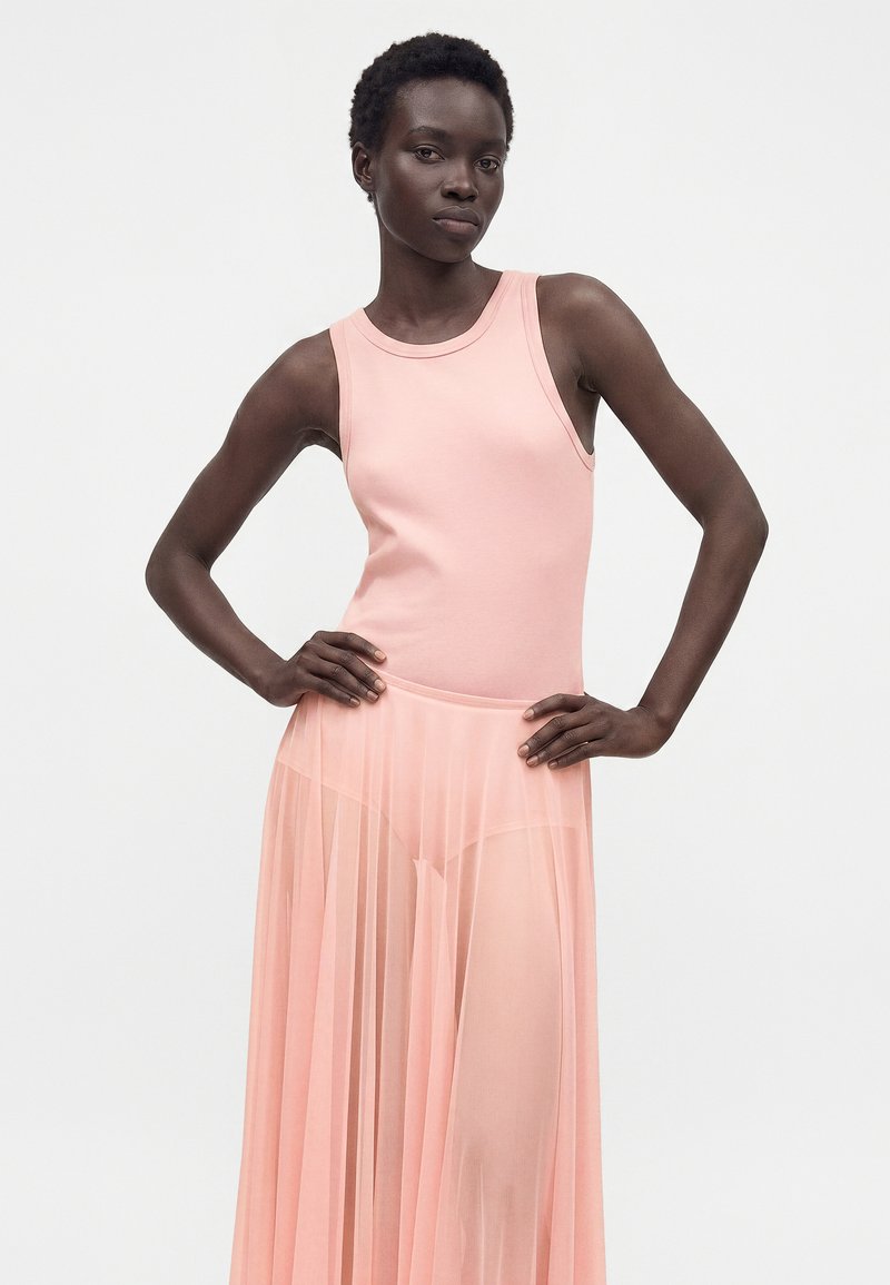 Femme portant un haut rose sans manches et une jupe plissée rose transparente, debout les mains sur les hanches devant un fond blanc uni.
