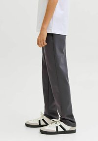 Personne debout portant une chemise blanche, un pantalon gris foncé et des baskets blanches avec des bandes noires sur un fond clair uni.