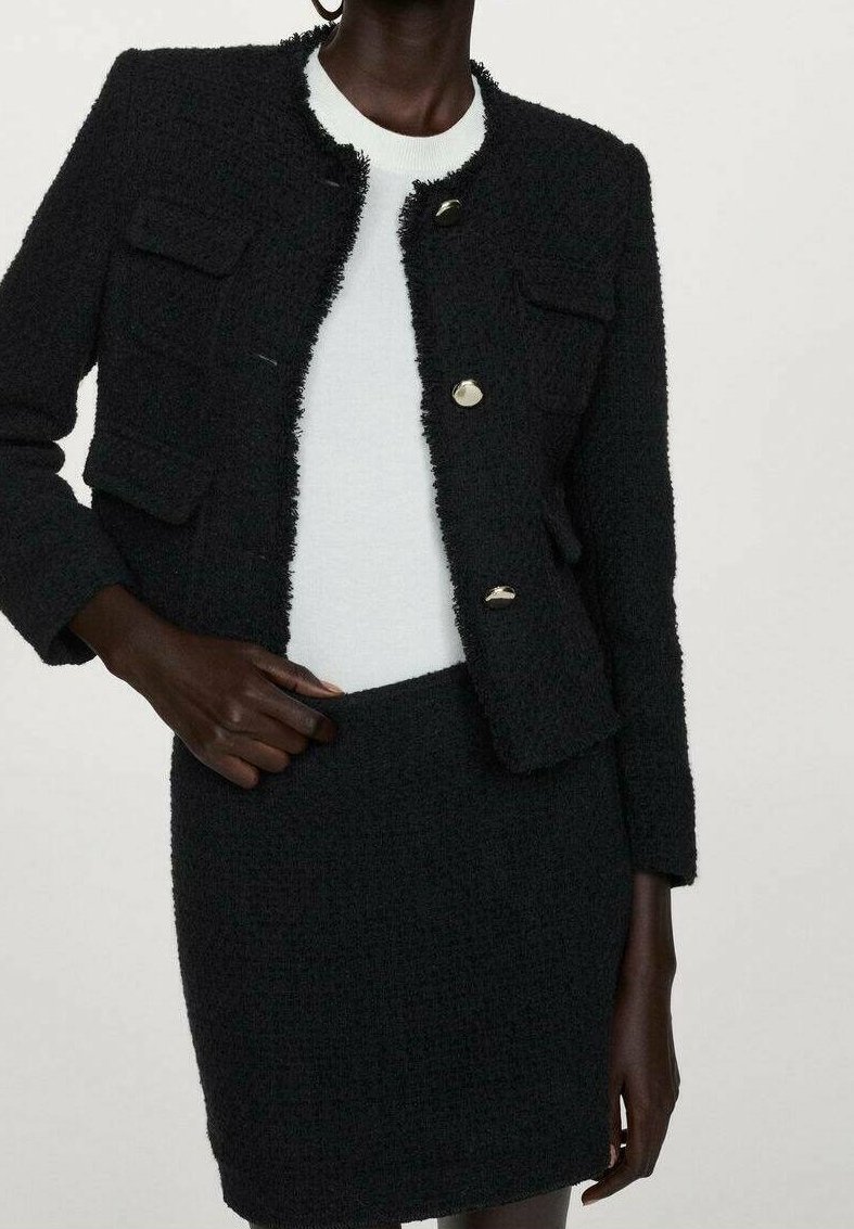 Veste noire texturée avec boutons dorés et jupe assortie portée sur un haut blanc, portée par une personne à la peau foncée sur un fond uni.