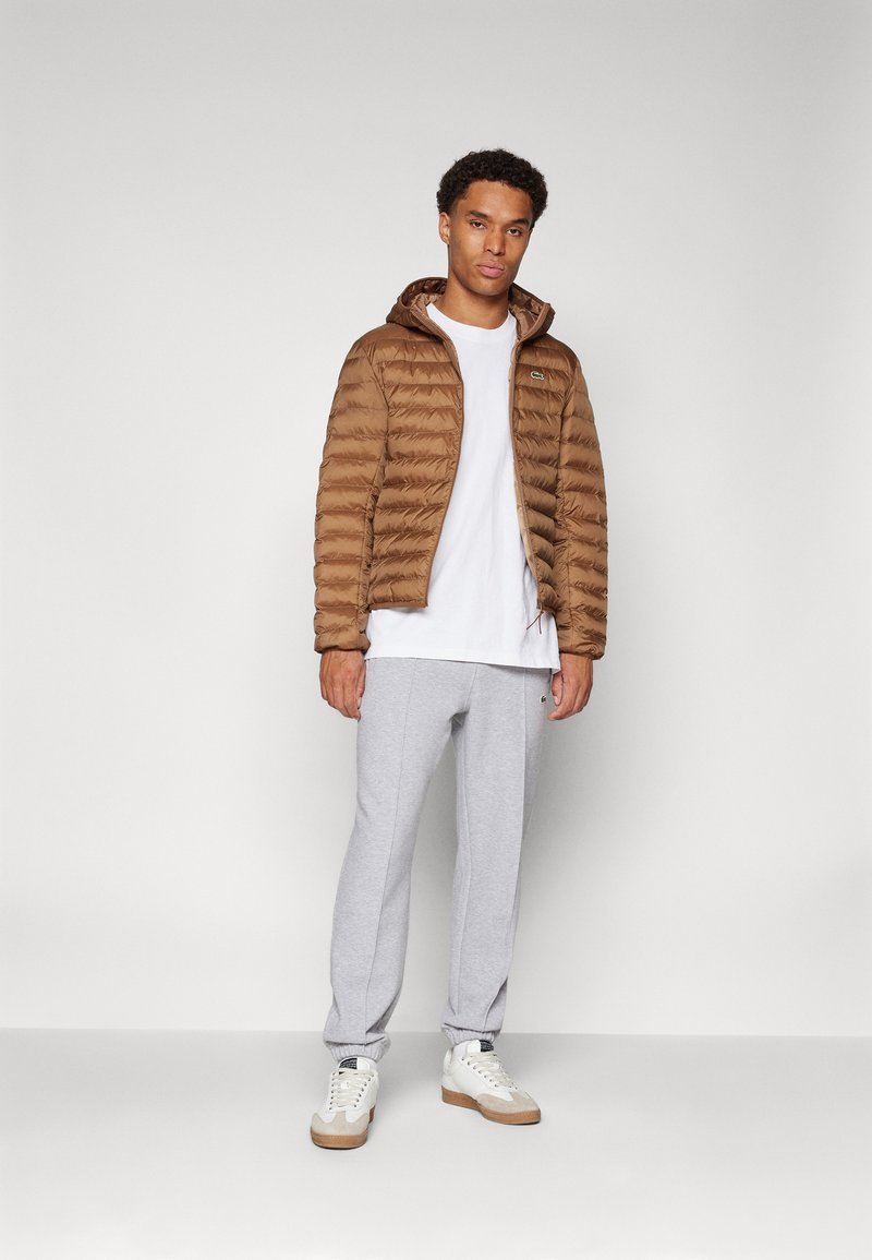 Giacca imbottita marrone con cappuccio, polsini a costine; t-shirt bianca sotto, pantaloni grigi e sneakers bianche con dettagli beige. Design semplice.