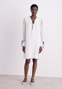 Weißes Kleid mit langen durchsichtigen Ärmeln, plissierter Textur und gerafftem Ausschnitt, wobei ein schwarzes Bindeband an der Vorderseite und kontrastierende Manschetten hervorgehoben werden.