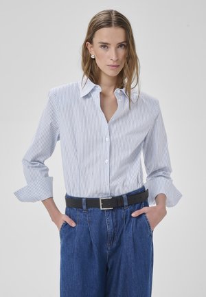Woman wearing a white pinstripe button-up shirt with rolled sleeves, blue high-waist jeans, black belt, and silver earrings, hands in pockets.