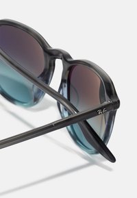 Ochelari de soare cu rame translucide gri, lentile cu un gradient de la teal la mov și brațe negre elegante cu un accent mic al logo-ului.