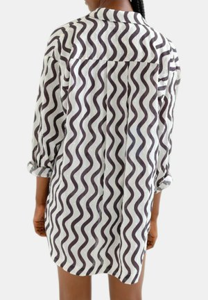 Personne portant une robe-chemise à manches longues avec des rayures ondulées noires sur fond blanc, manches retroussées, vue de dos sur un fond uni.