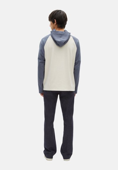Felpa con cappuccio dal corpo crema, maniche e cappuccio blu. Tessuto morbido, vestibilità casual. Abbinata a pantaloni blu scuro e scarpe chiare.