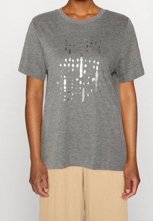 Person iført en grå kortærmet T-shirt med sølvfarvet abstrakt prikket design og beige ribstrikkede bukser, stående mod en ensfarvet baggrund.